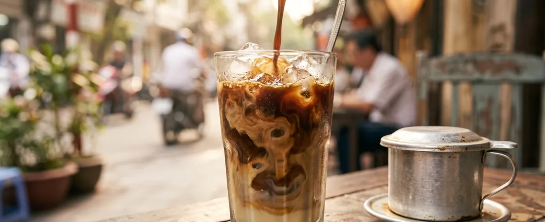 A frosty glass of cà phê sữa đá (Vietnamese iced milk coffee) — dark coffee poured over ice with a swirl of sweetened condensed milk, a metal phin filter beside it on a small street-café table.